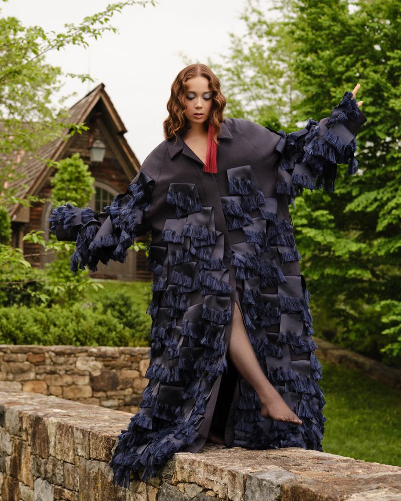 Fashionable woman strikes a pose outdoors in a long, dark blue coat with fringe details, complemented by trees and a charming house.
