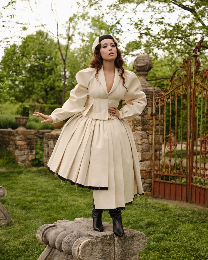 Adult female in an oversized cream-colored outfit with a matching hat and black boots standing on a stone pedestal in a garden.