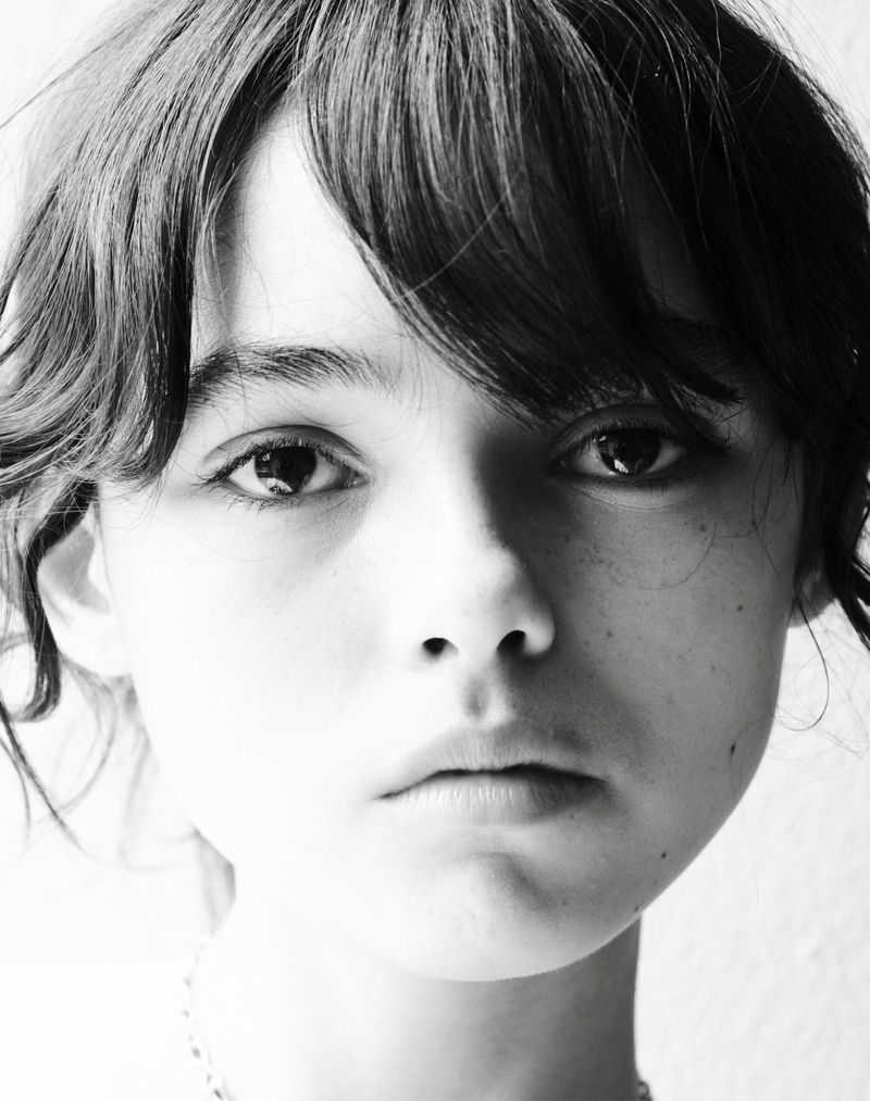 Close-up portrait of a young person with dark, curly hair, gazing directly forward in a black and white photograph with a bright background.