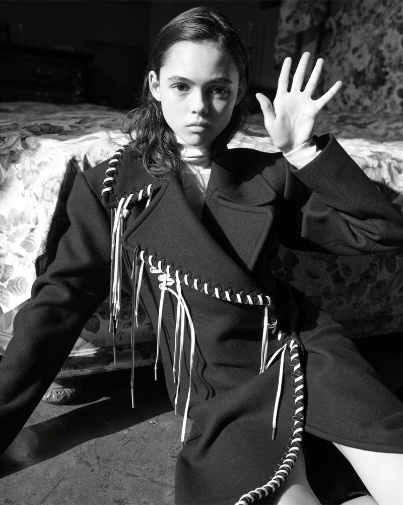 Black and white portrait of a young person in a dark coat with a light turtleneck, sitting in a room with a patterned bed.
