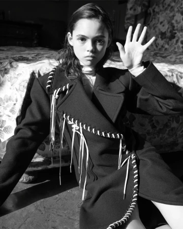 Black and white portrait of a young person in a dark coat with a light turtleneck, sitting in a room with a patterned bed.
