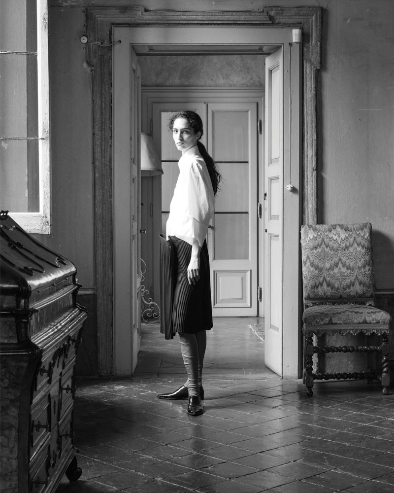Model in a white collared shirt and dark pleated skirt standing in an open stone interior doorway in black and white.