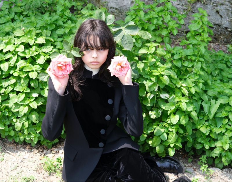 Young woman in a black suit holds pink roses in an outdoor setting with greenery and natural light, creating a contrast between formal wear and nature.