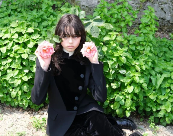 Young woman in a black suit holds pink roses in an outdoor setting with greenery and natural light, creating a contrast between formal wear and nature.