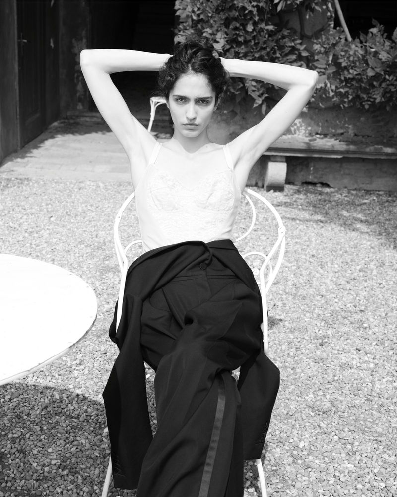 Black and white portrait of a woman with her hands behind her head, sitting on a chair outdoors with a jacket draped over it.