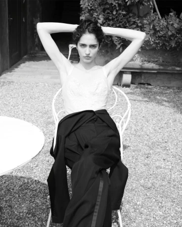 Black and white portrait of a woman with her hands behind her head, sitting on a chair outdoors with a jacket draped over it.