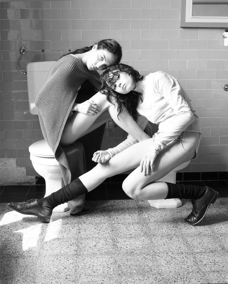 Black and white photo of two women in a tiled bathroom, one sitting on the toilet, the other leaning against her, wearing casual clothing and boots.