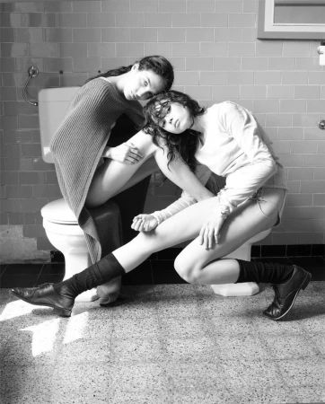 Black and white photo of two women in a tiled bathroom, one sitting on the toilet, the other leaning against her, wearing casual clothing and boots.