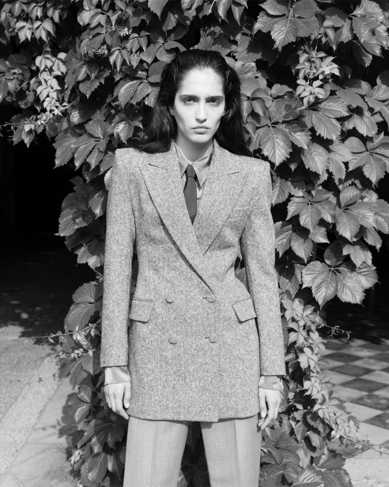 Black and white portrait of a woman in a double-breasted blazer and slacks standing in front of ivy leaves in a studio setting.