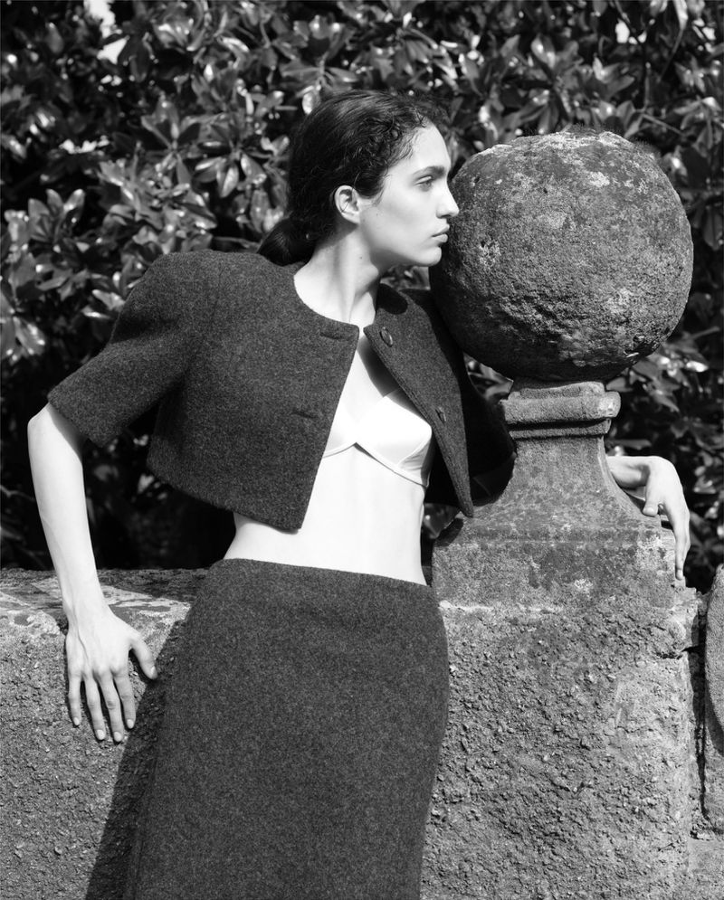 Monochrome portrait of a young woman in a skirt and jacket posing outdoors by stone architectural element with green foliage backdrop.
