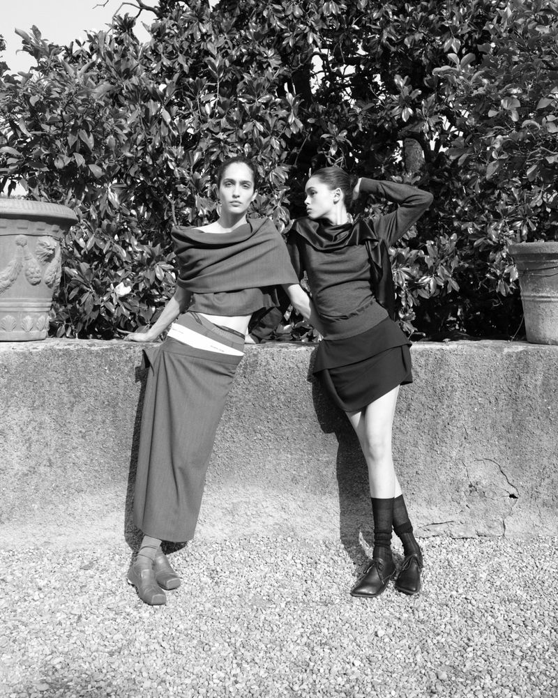 Two fashion models pose outdoors in draped clothing against a garden wall backdrop in a black and white fashion photograph.