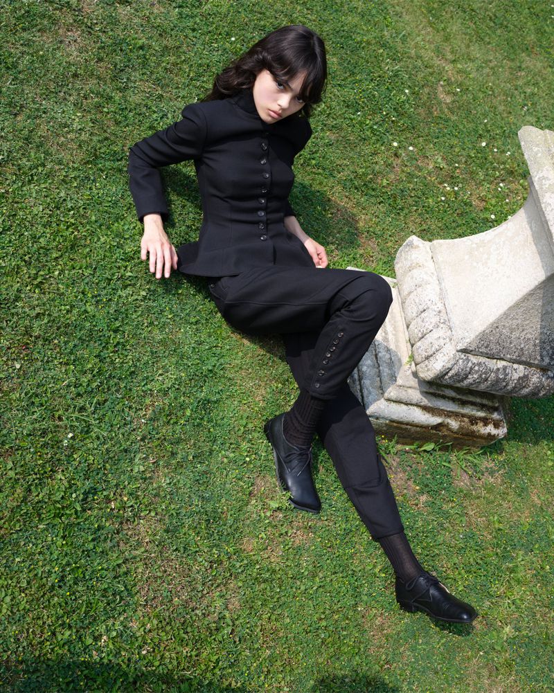 Woman in black suit lies on green grass next to a stone structure, captured in a full shot on a sunny day.