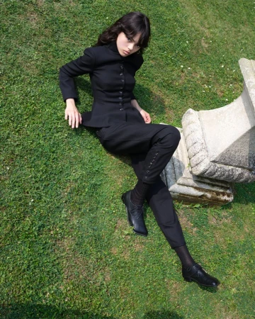 Woman in black suit lies on green grass next to a stone structure, captured in a full shot on a sunny day.