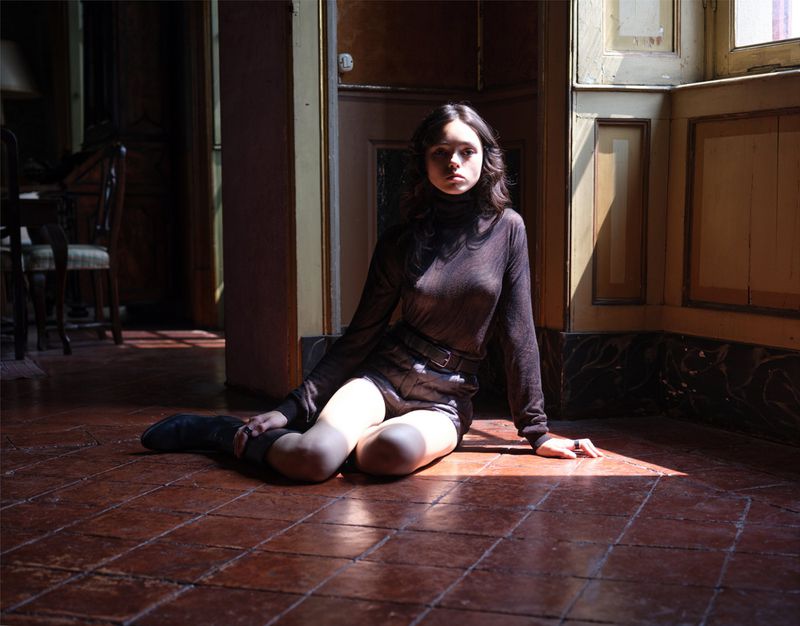 Young woman with long brown hair and black top and shorts poses in room with red-tiled floor and soft window light.