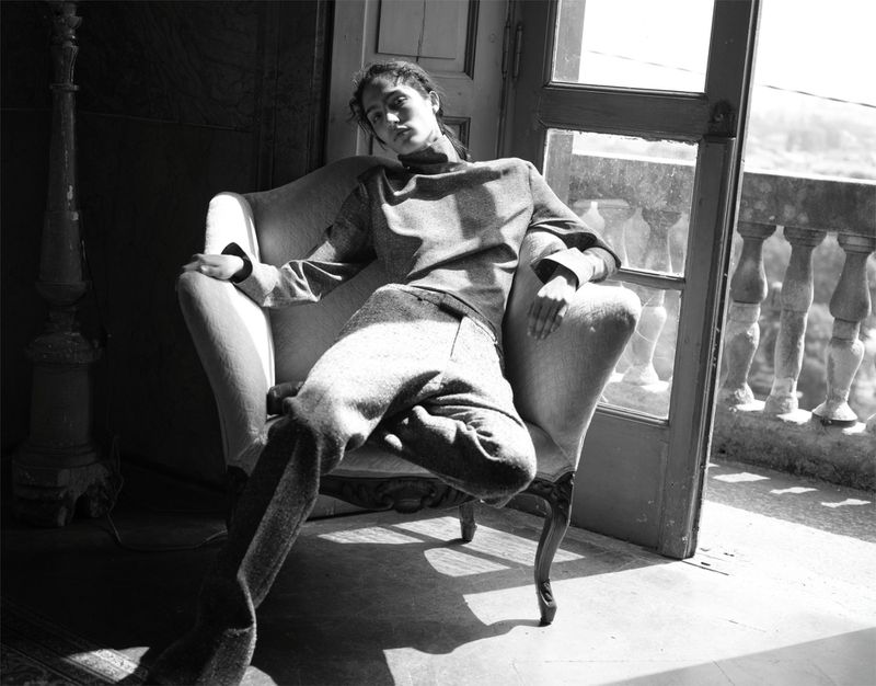 An adult female sits in a chair in a room with sunlight shining through a doorway leading to a balcony in a monochrome photograph.