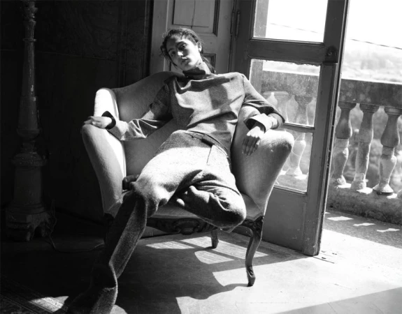 An adult female sits in a chair in a room with sunlight shining through a doorway leading to a balcony in a monochrome photograph.