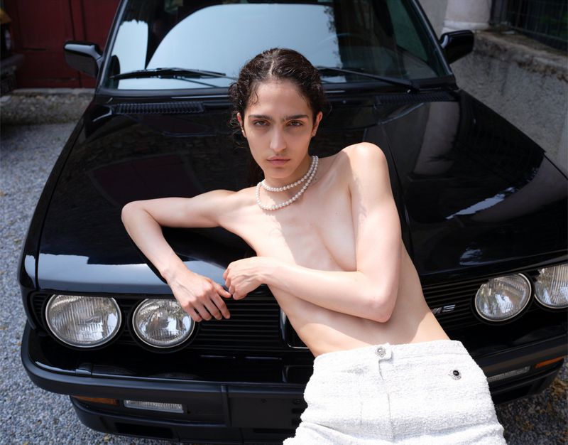 Adult female poses with a classic black car in an outdoor setting, wearing a pearl necklace and white bottoms in natural light.