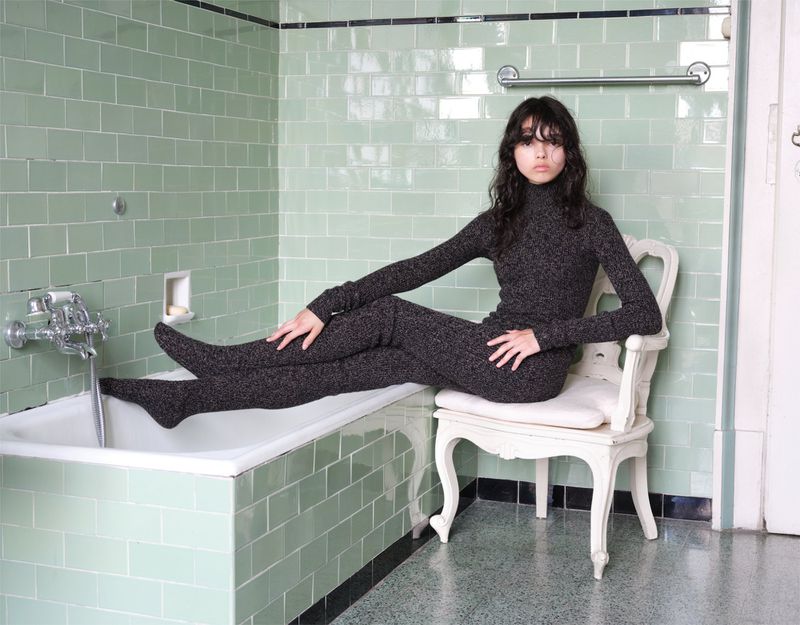 Young woman in a grey knit set poses on a white chair in a tiled bathroom. She sits with legs extended, looking directly at the camera.