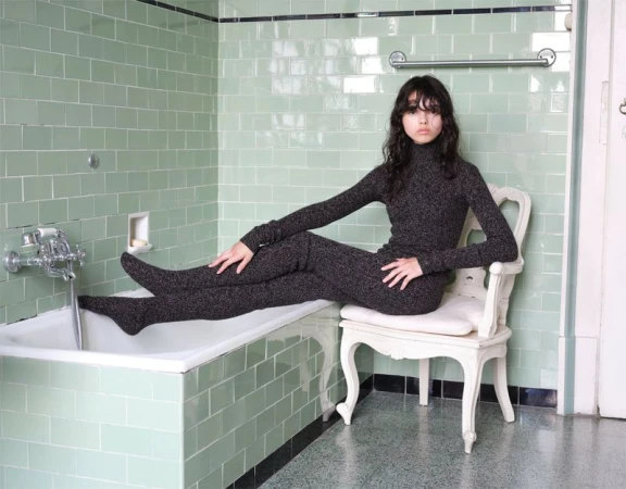 Young woman in a grey knit set poses on a white chair in a tiled bathroom. She sits with legs extended, looking directly at the camera.