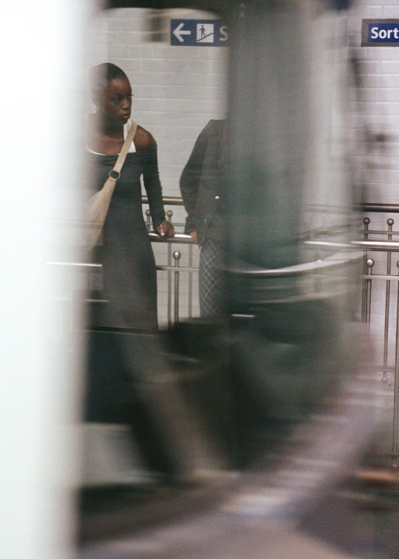 Woman in a black dress waits near a metro platform with a shoulder bag in an underground station with blurred motion, with another figure.
