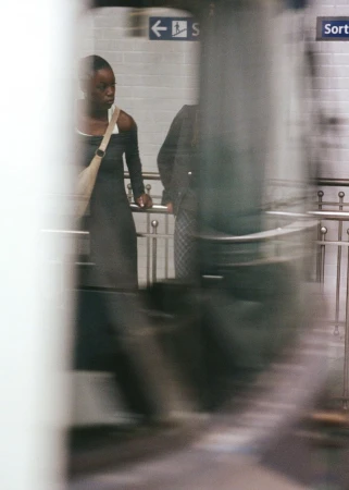 Woman in a black dress waits near a metro platform with a shoulder bag in an underground station with blurred motion, with another figure.