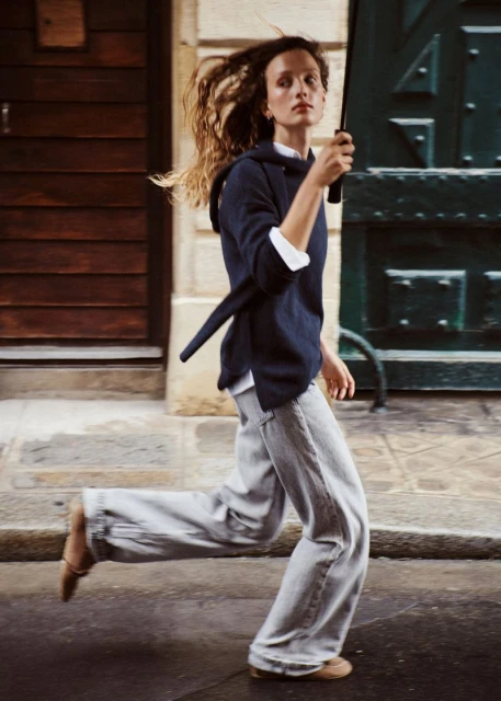 A woman runs on a city street wearing a navy hooded sweater, wide-leg denim pants, and flats. She's holding a black umbrella.