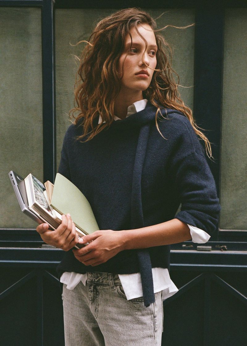 A young adult woman poses indoors, wearing a navy sweater, white collared shirt, and grey jeans, holding books near a window.