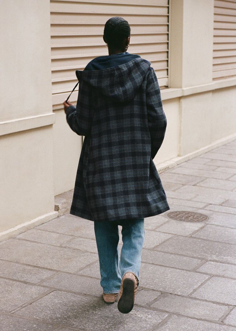 Back view of an adult woman walking on a sidewalk wearing a blue and grey plaid coat, jeans, and tan shoes on a sunny day.