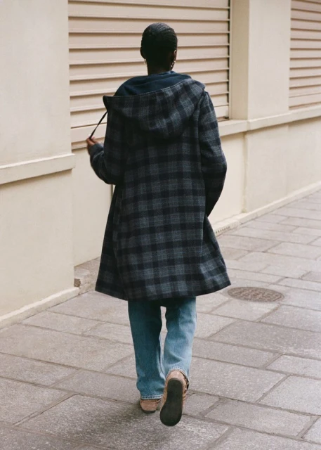 Back view of an adult woman walking on a sidewalk wearing a blue and grey plaid coat, jeans, and tan shoes on a sunny day.