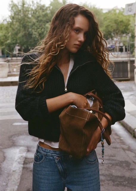 A young woman with long curly hair poses outdoors with a brown suede bag, wearing a black jacket and blue jeans. Street scene background.