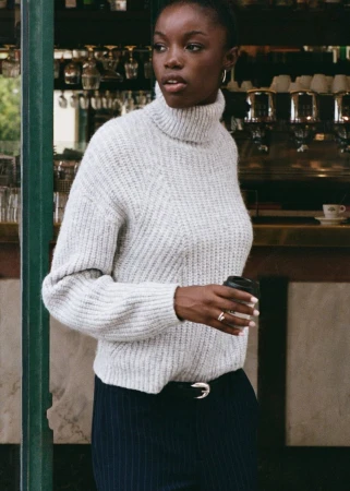 Portrait of a young Black woman in a grey turtleneck sweater holding a coffee cup in an urban cafe setting with muted natural light.