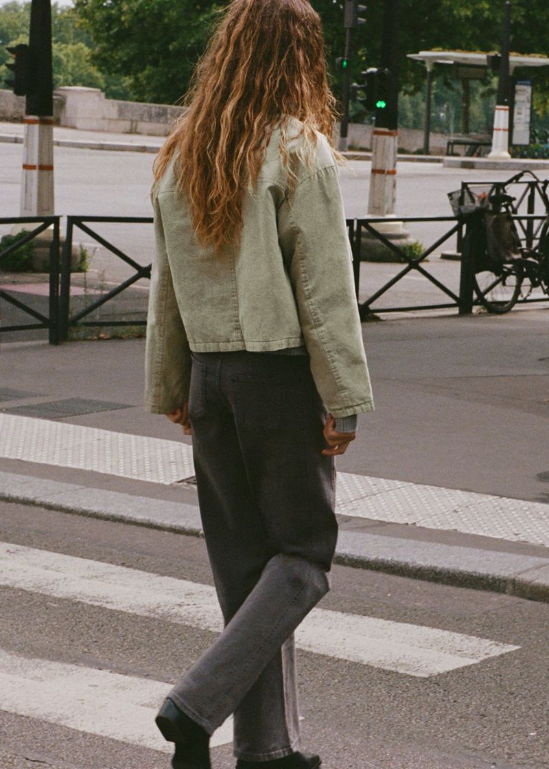 Back view of adult female with long wavy hair crossing a street on a crosswalk wearing a light green jacket and dark jeans in an urban area.