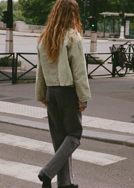 Back view of adult female with long wavy hair crossing a street on a crosswalk wearing a light green jacket and dark jeans in an urban area.