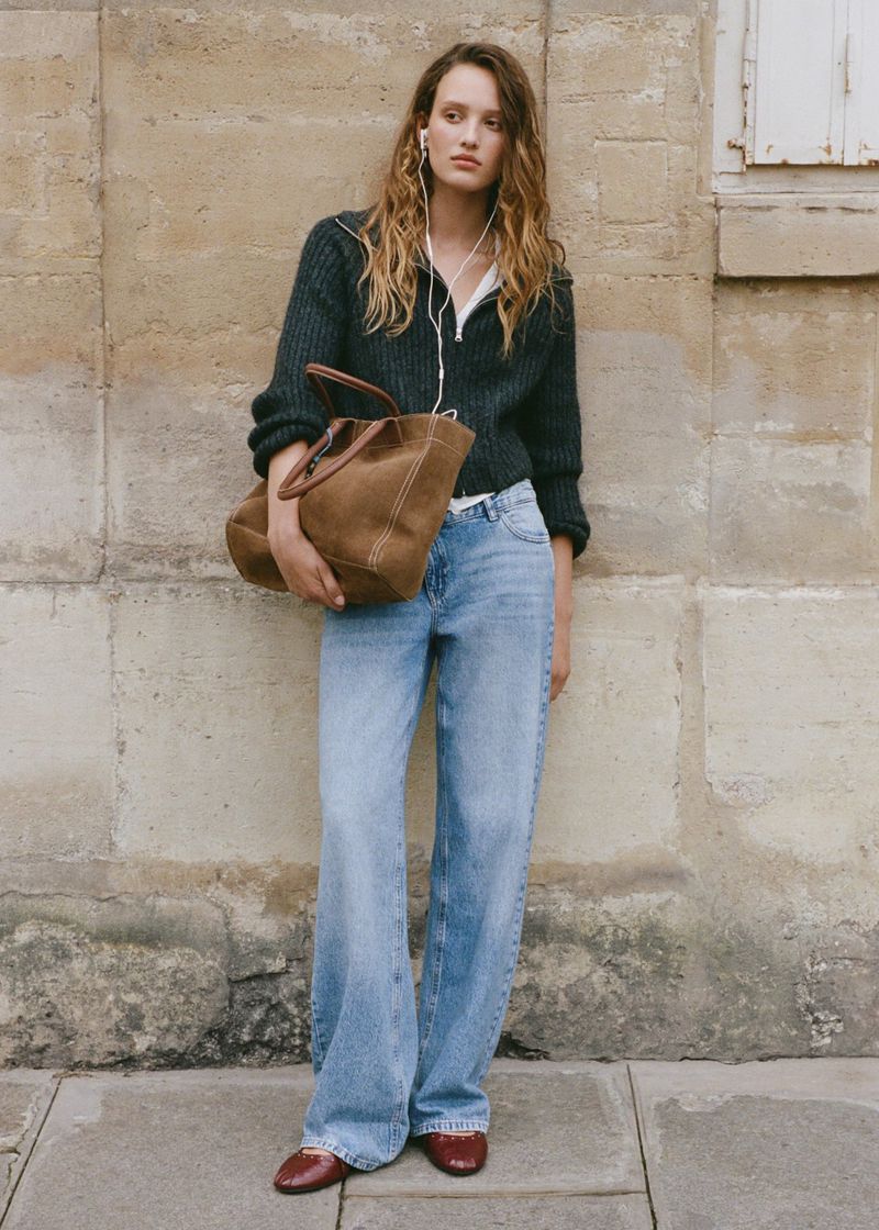 A young adult woman stands in a full shot wearing wide leg jeans, a dark collared cardigan, carrying a brown tote bag, and wearing flats.