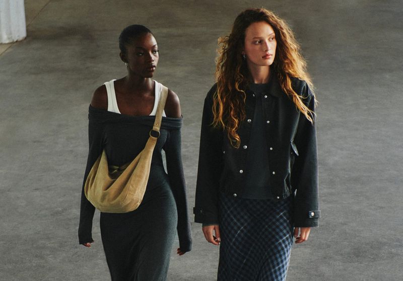 Two adult women in fashionable apparel walk in an open indoor area. One is wearing an olive-green bag on her shoulder.