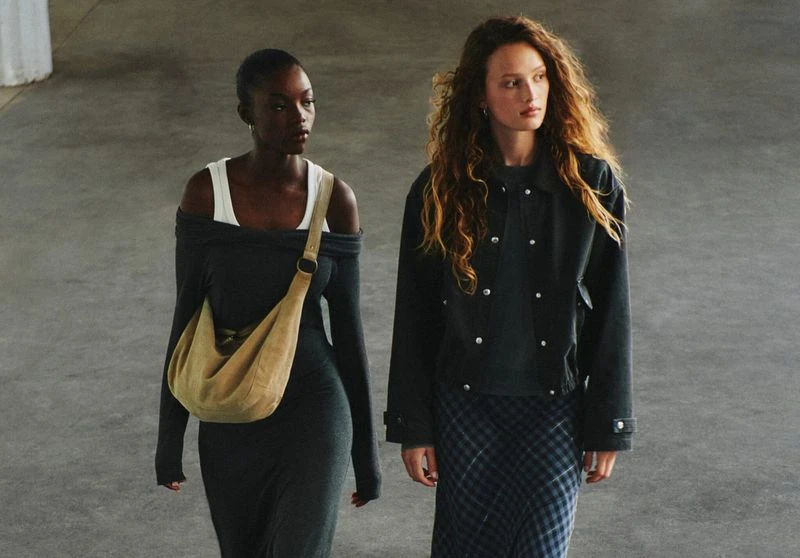 Two adult women in fashionable apparel walk in an open indoor area. One is wearing an olive-green bag on her shoulder.