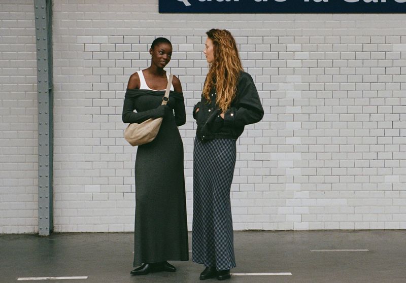Two women in stylish dresses stand in a subway station, one in green, the other in a jacket and checked dress, by tiled walls.