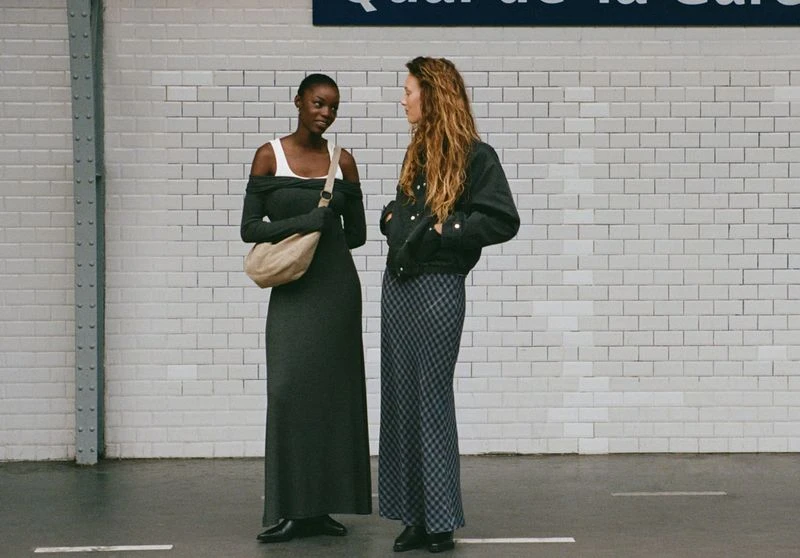 Two women in stylish dresses stand in a subway station, one in green, the other in a jacket and checked dress, by tiled walls.