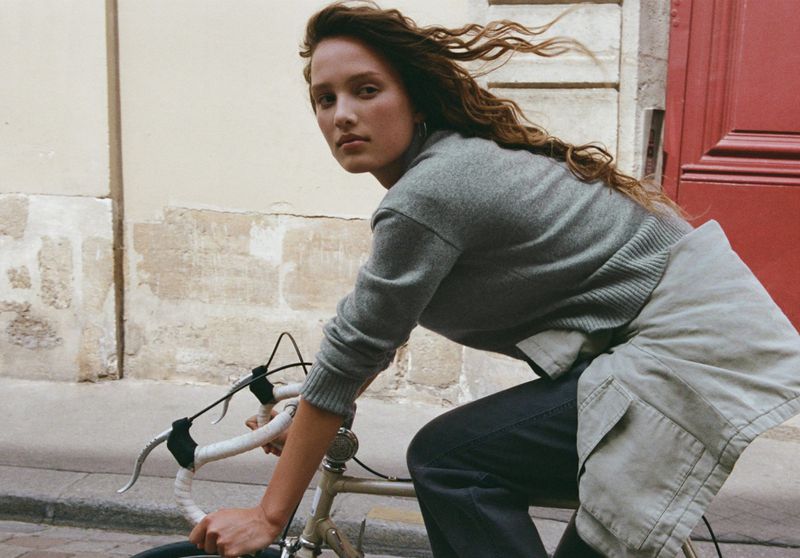 Woman rides vintage bicycle outdoors, wearing gray sweater and dark pants, with weathered stone wall and red door background.