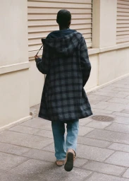 Rear view of a person walking along a paved sidewalk wearing a dark plaid hooded coat, blue jeans, and tan shoes.