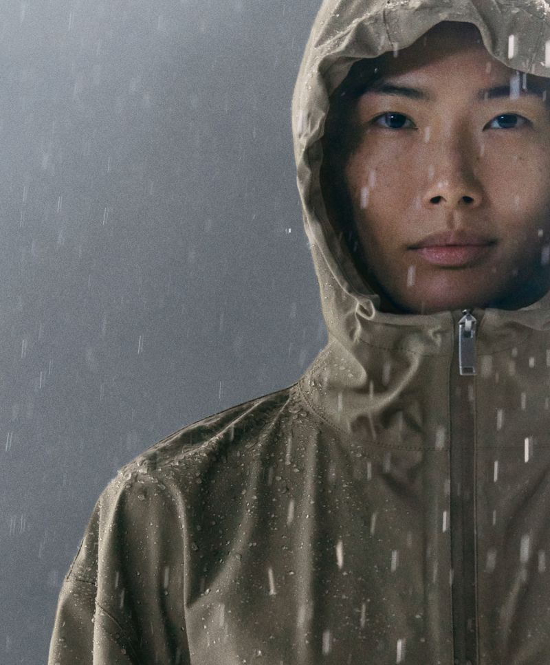Person in a hooded tan raincoat stands in the rain. The photo is a close-up shot with a grey background and natural light.