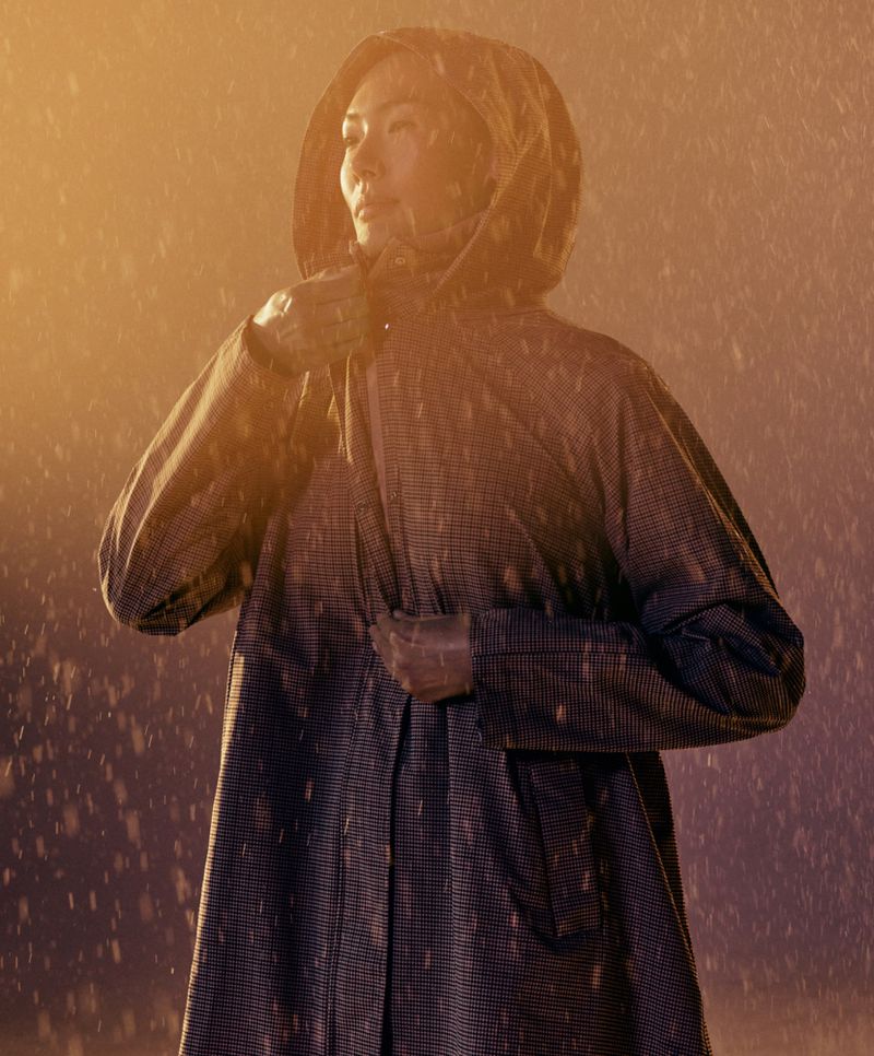 An Asian woman in a hooded raincoat stands in the rain, looking off to the side with a slightly furrowed brow, illuminated by warm light.