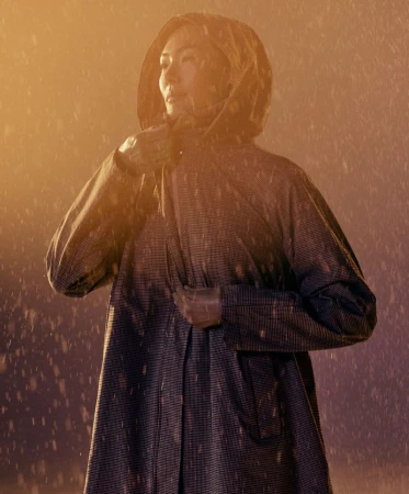 An Asian woman in a hooded raincoat stands in the rain, looking off to the side with a slightly furrowed brow, illuminated by warm light.