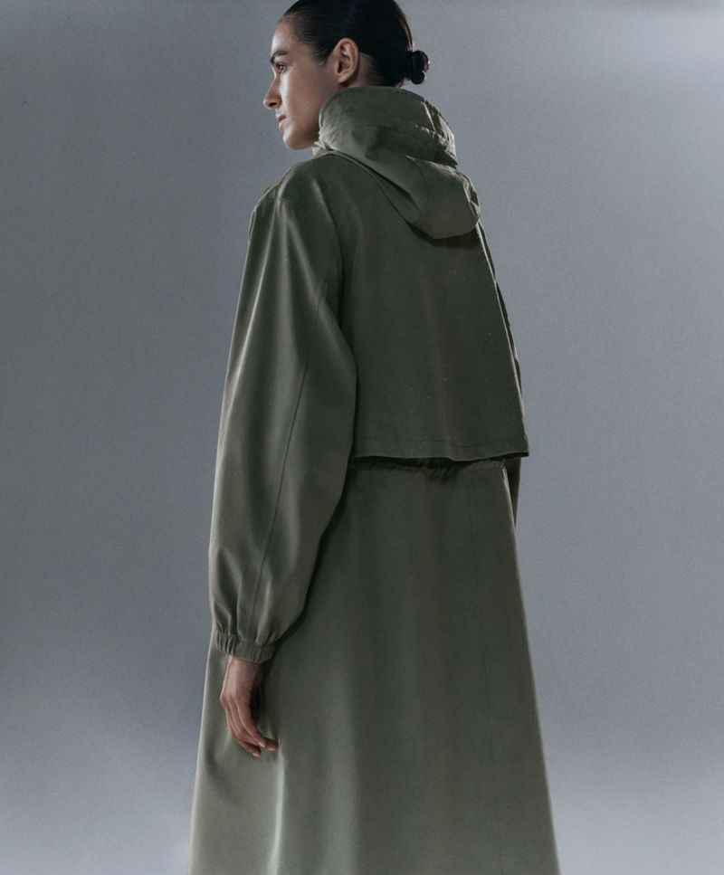 An adult female model poses in a long, olive green hooded raincoat in a studio setting. She's shot from the back at medium distance.