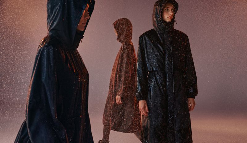 Three women in hooded raincoats stand in the rain, creating a moody, fashion-forward image with cool tones and a sense of protection.