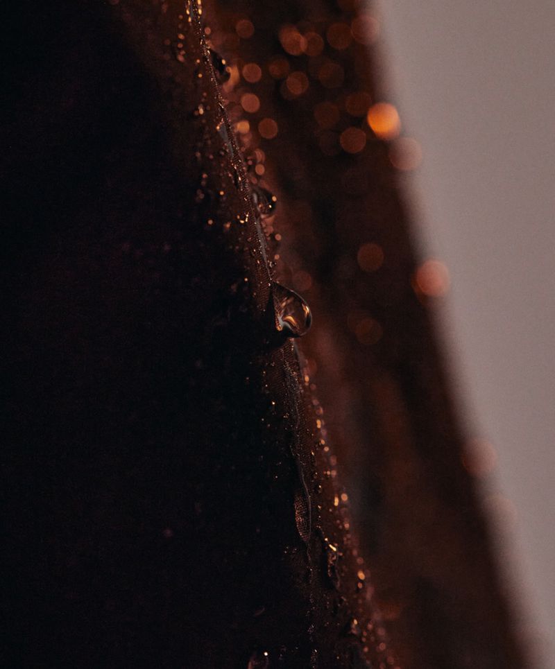 Macro shot of water droplets on a dark, textured surface with blurred golden highlights in background, creating moody, wet aesthetic.