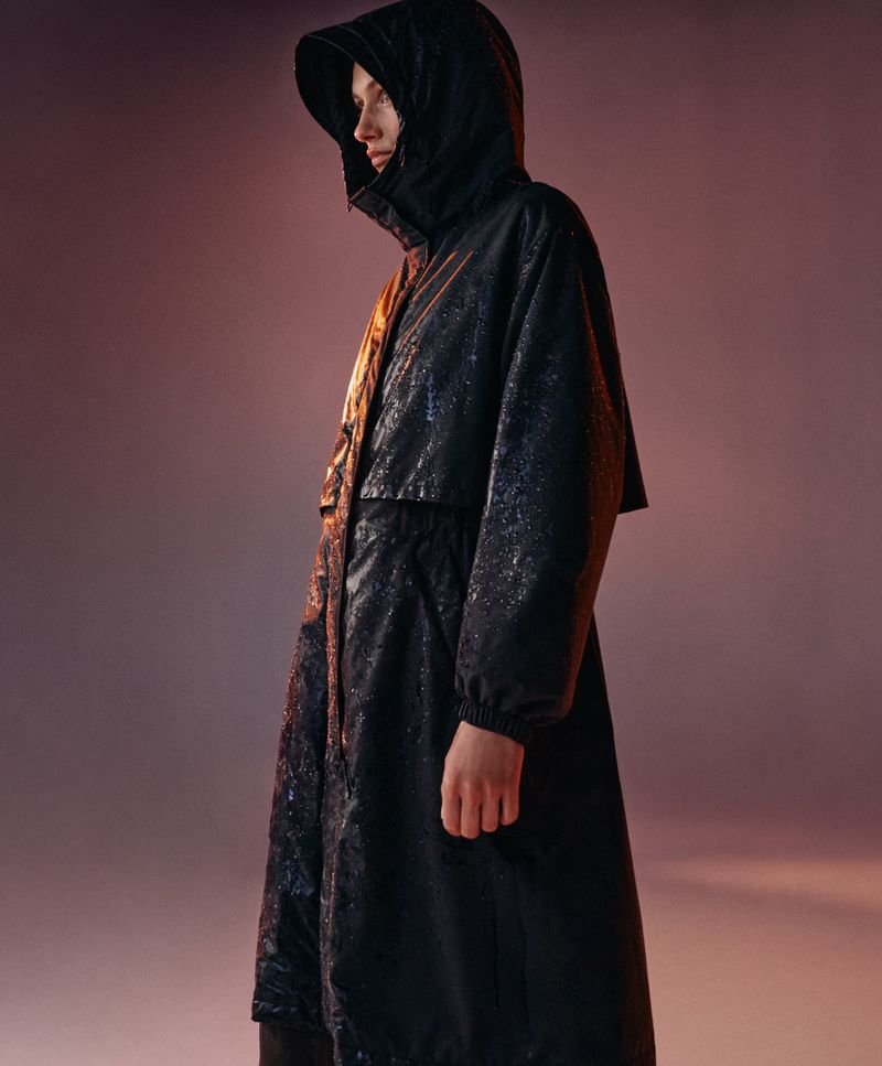 A woman in a black, shiny, hooded raincoat stands in a studio with diffused lighting and a blend of orange and brown hues.