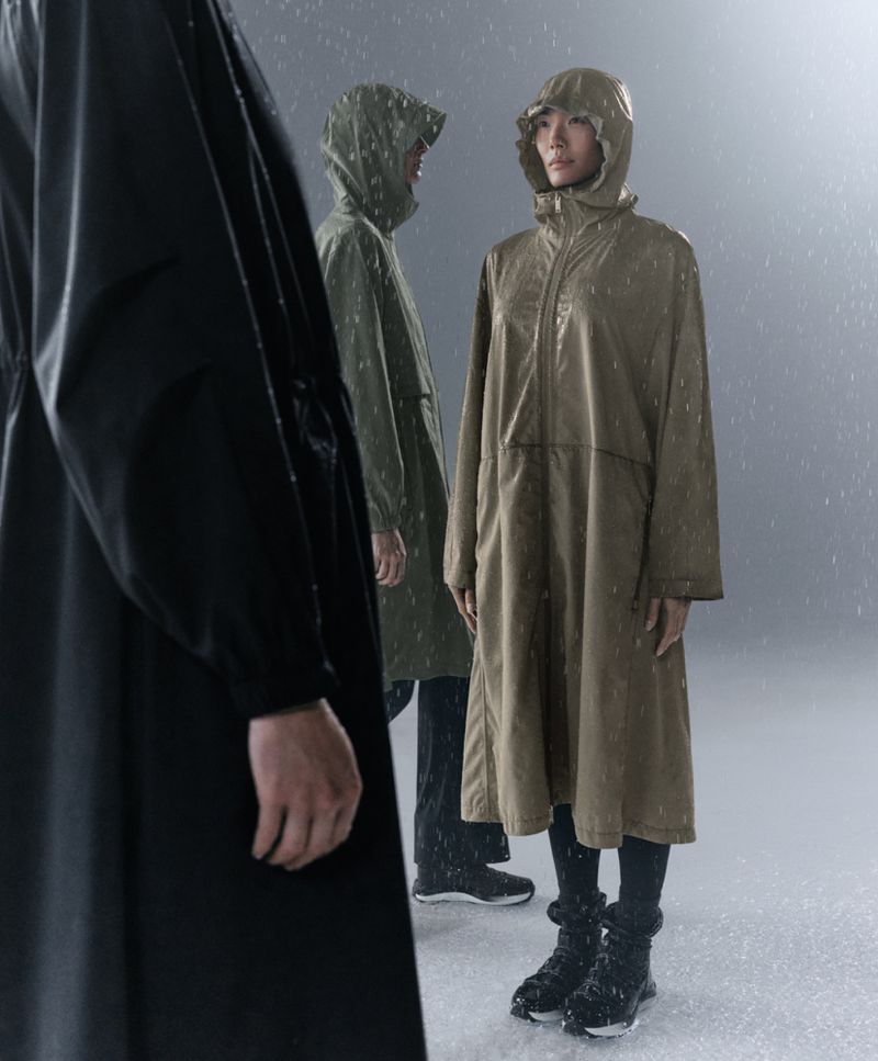Three people stand in the rain wearing long hooded coats. The central figure is an Asian woman looking ahead, wearing a tan rain coat.