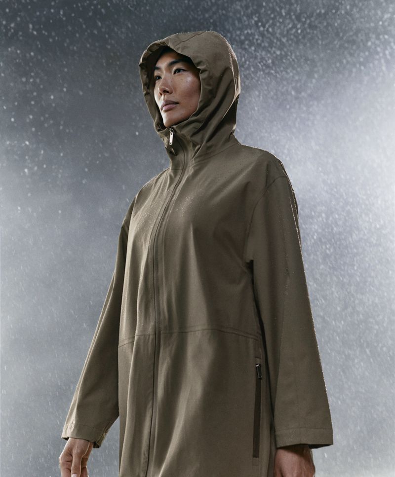 An Asian woman in a khaki hooded raincoat stands in the rain, captured in a medium shot. The scene is lit from the side.