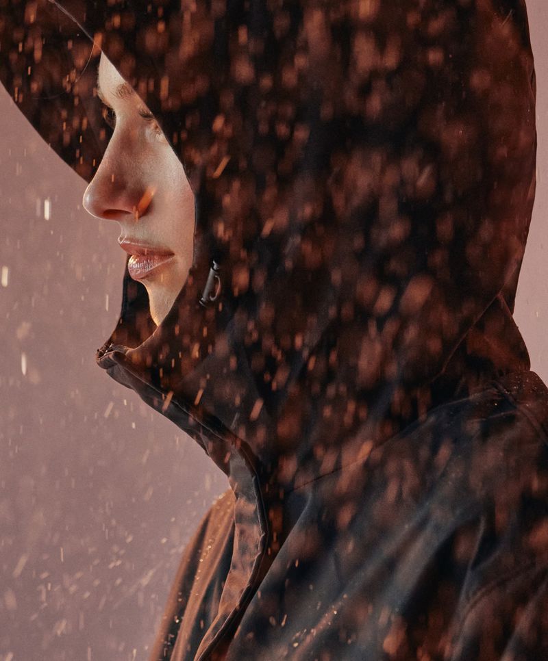 Profile of an adult in a hooded jacket, seen in a close-up shot with an orange-toned color grade and light sprinkling. Fashion and beauty.